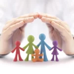 Dłonie tworzące ochronny gest nad kolorowymi figurkami przedstawiającymi rodzinę z dzieckiem.