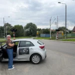 Instruktorka OSK Tailor Iza w okularach stojąca ze skrzyżowanymi rękami przy otwartych drzwiach srebrnego samochodu nauki jazdy (Kia Rio 4)z logo "TAILOR Ośrodek Szkolenia Kierowców". W tle widoczny jest przejazd kolejowy.